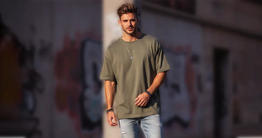 A stylish man wears an oversized olive t-shirt and ripped jeans outdoors.