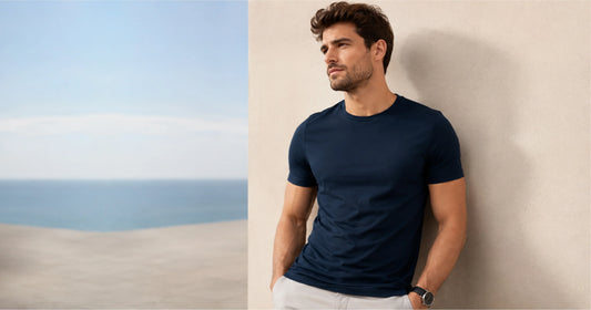 Man wearing a fitted navy T-shirt and light trousers beside a seaside wall.