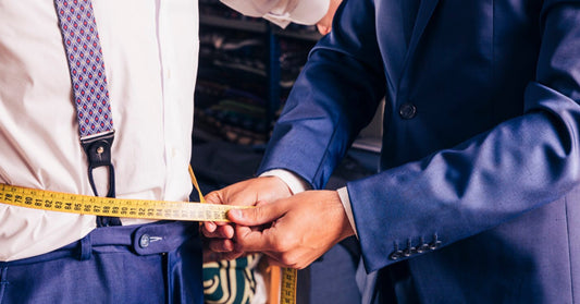 A tailor uses a yellow tape to measure the waist of a man wearing dress pants and suspenders.